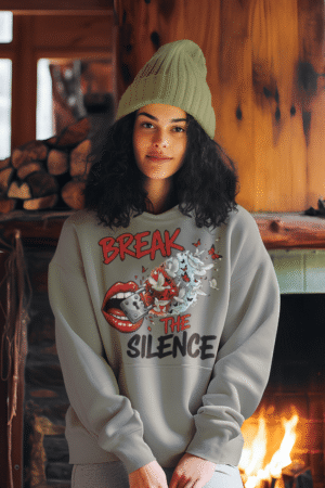 A young woman, possibly of Caucasian descent, with light brown hair pulled back, wearing a grey PodLyft Women's Break the Silence Hoodie. The hoodie features a graphic of a padlocked mouth with doves and butterflies bursting out, and the text "BREAK THE SILENCE" and "RAISE YOUR VOICE." She is looking directly at the camera with a confident expression, standing against a plain light grey background.