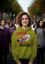 A young woman, possibly of Caucasian descent, with light brown hair pulled back, wearing an olive green PodLyft Women's Break the Silence Hoodie. The hoodie features a graphic of a padlocked mouth with doves and butterflies bursting out, and the text "BREAK THE SILENCE" and "RAISE YOUR VOICE." She is looking directly at the camera with a confident expression, standing against a plain light grey background.