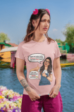 A woman wearing a baby pink PodLyft Girls Savage Queen Tee with a funny graphic and Hindi text. She is standing outdoors with a lake and mountains in the background. The graphic features a woman giving a thumbs-up, with text bubbles that translate to "If a guy proposes to me, I reject him" and "But I salute his choice.