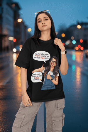 A woman wearing a black PodLyft Girls Savage Queen Tee with a funny graphic and Hindi text. She is standing outdoors with a blurred street and city lights in the background. The graphic features a woman giving a thumbs-up, with text bubbles that translate to "If a guy proposes to me, I reject him" and "But I salute his choice.