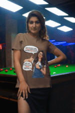 A woman wearing a coffee brown PodLyft Girls Savage Queen Tee with a funny graphic and Hindi text. She is standing indoors next to a pool table. The graphic features a woman giving a thumbs-up, with text bubbles that translate to "If a guy proposes to me, I reject him" and "But I salute his choice.