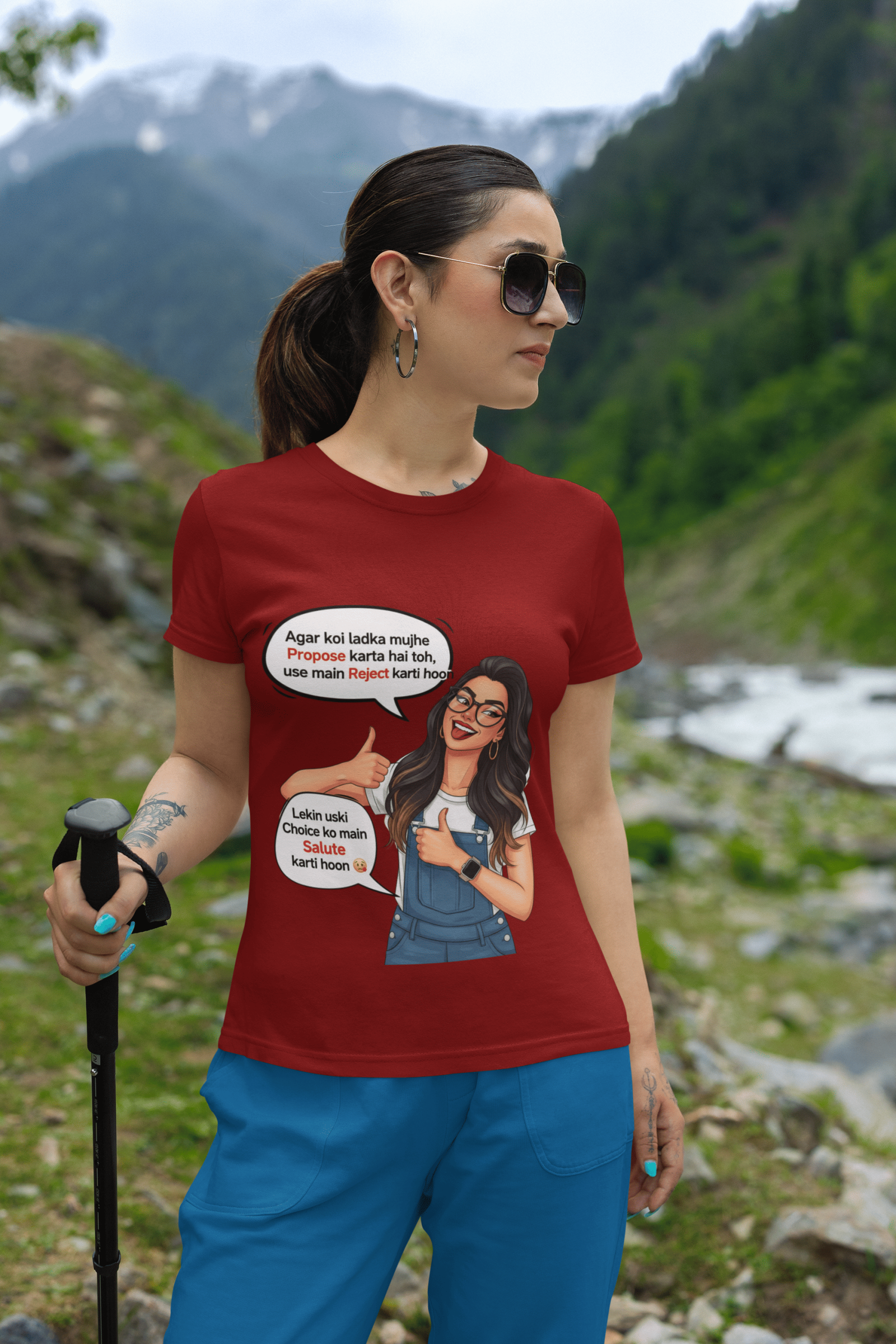 A woman wearing a maroon PodLyft Girls Savage Queen Tee with a funny graphic and Hindi text. She is standing outdoors with a mountain and river background, holding a hiking pole. The graphic features a woman giving a thumbs-up, with text bubbles that translate to "If a guy proposes to me, I reject him" and "But I salute his choice.