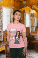 A woman wearing a pink PodLyft Girls Savage Queen Tee with a funny graphic and Hindi text. She is standing indoors in a cozy, wood-paneled room. The graphic features a woman giving a thumbs-up, with text bubbles that translate to "If a guy proposes to me, I reject him" and "But I salute his choice.