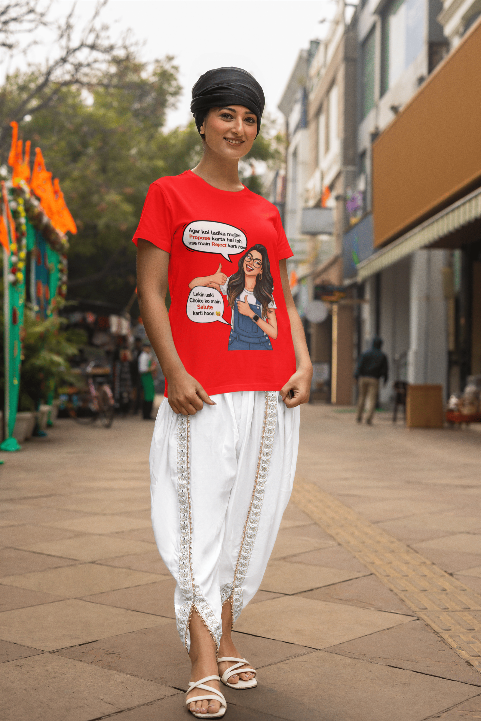 A woman smiling while wearing a red PodLyft Girls Savage Queen Tee and white traditional Indian clothing. She is standing on a city street. The graphic features a woman giving a thumbs-up, with Hindi text bubbles that translate to "If a guy proposes to me, I reject him" and "But I salute his choice.