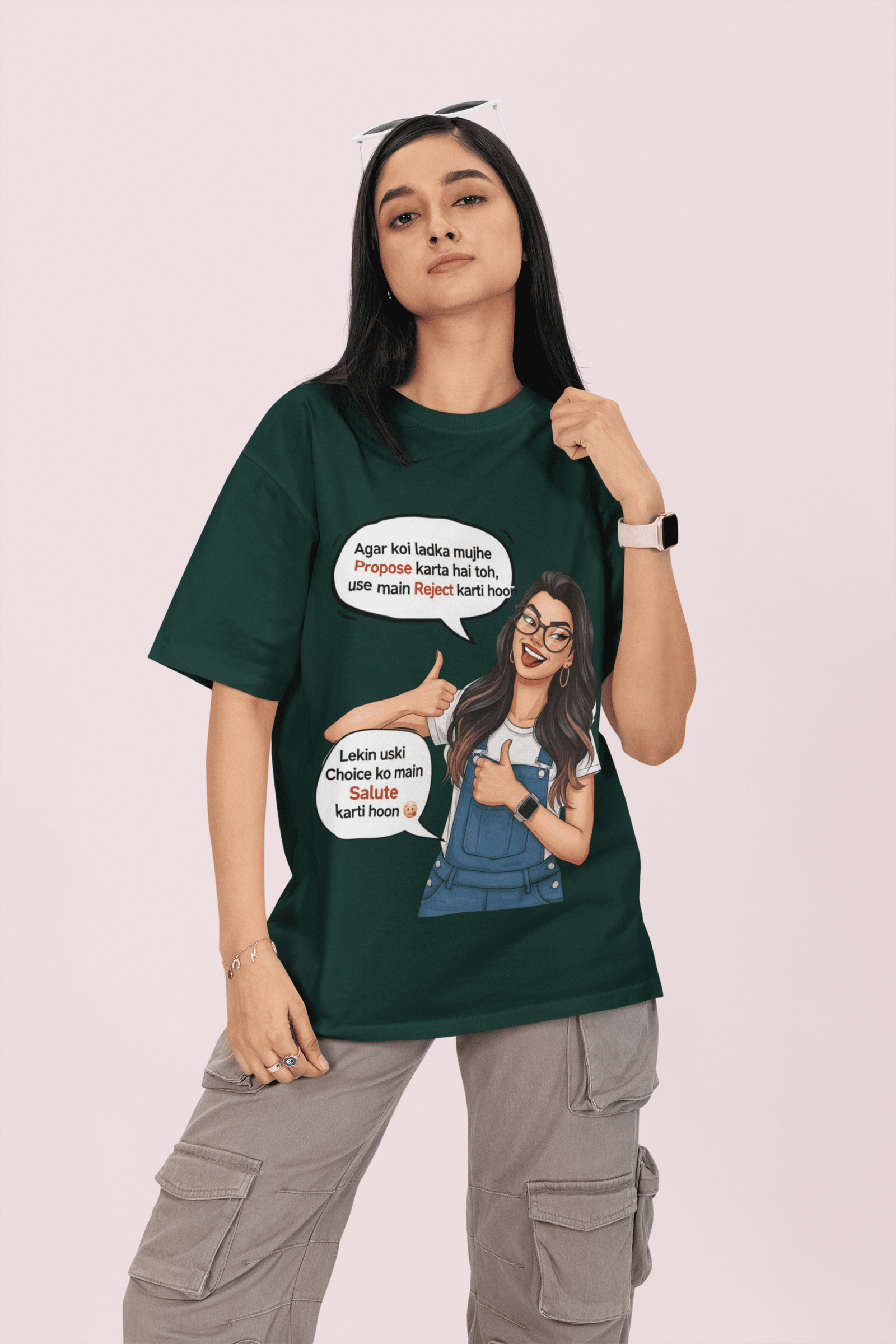 A woman wearing a dark green PodLyft Girls Savage Queen Tee with a funny, relatable graphic. She is standing against a light pink background, posing with one hand on her hair. The graphic features a stylized cartoon woman giving a thumbs-up, with two Hindi text bubbles that translate to "If a guy proposes to me, I reject him" and "But I salute his choice.