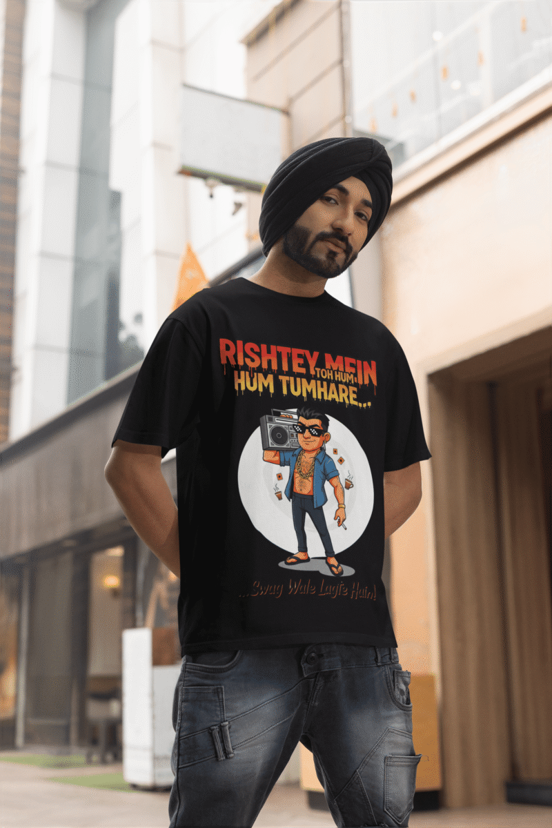 A young man, possibly of Caucasian descent, with short dark hair and a beard, wearing a black PodLyft Men's Bollywood Swag T-shirt. The t-shirt features a graphic with a funny movie dialogue. He is looking directly at the camera with a confident expression, standing against a plain light grey background.