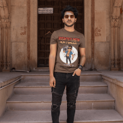 A young man, possibly of Caucasian descent, with short dark hair and a beard, wearing a coffee brown PodLyft Men's Bollywood Swag T-shirt. The t-shirt features a graphic with a funny movie dialogue. He is looking directly at the camera with a confident expression, standing against a plain light grey background.