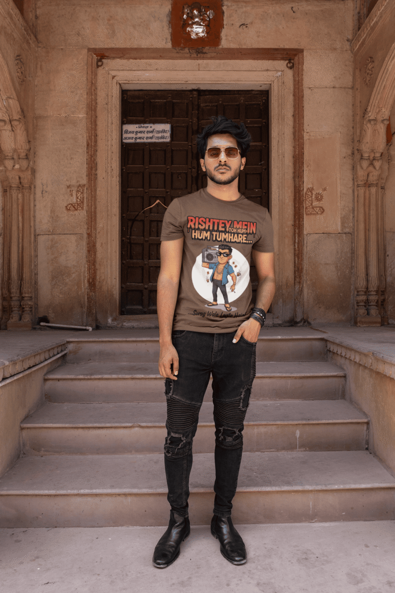 A young man, possibly of Caucasian descent, with short dark hair and a beard, wearing a coffee brown PodLyft Men's Bollywood Swag T-shirt. The t-shirt features a graphic with a funny movie dialogue. He is looking directly at the camera with a confident expression, standing against a plain light grey background.