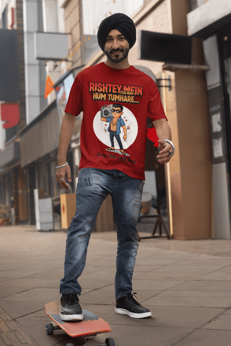 A young man, possibly of Caucasian descent, with short dark hair and a beard, wearing a maroon PodLyft Men's Bollywood Swag T-shirt. The t-shirt features a graphic with a funny movie dialogue. He is looking directly at the camera with a confident expression, standing against a plain light grey background.
