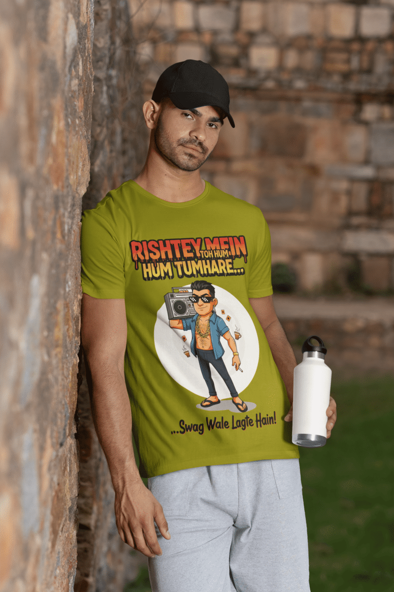 A young man, possibly of Caucasian descent, with short dark hair and a beard, wearing an olive green PodLyft Men's Bollywood Swag T-shirt. The t-shirt features a graphic with a funny movie dialogue. He is looking directly at the camera with a confident expression, standing against a plain light grey background.