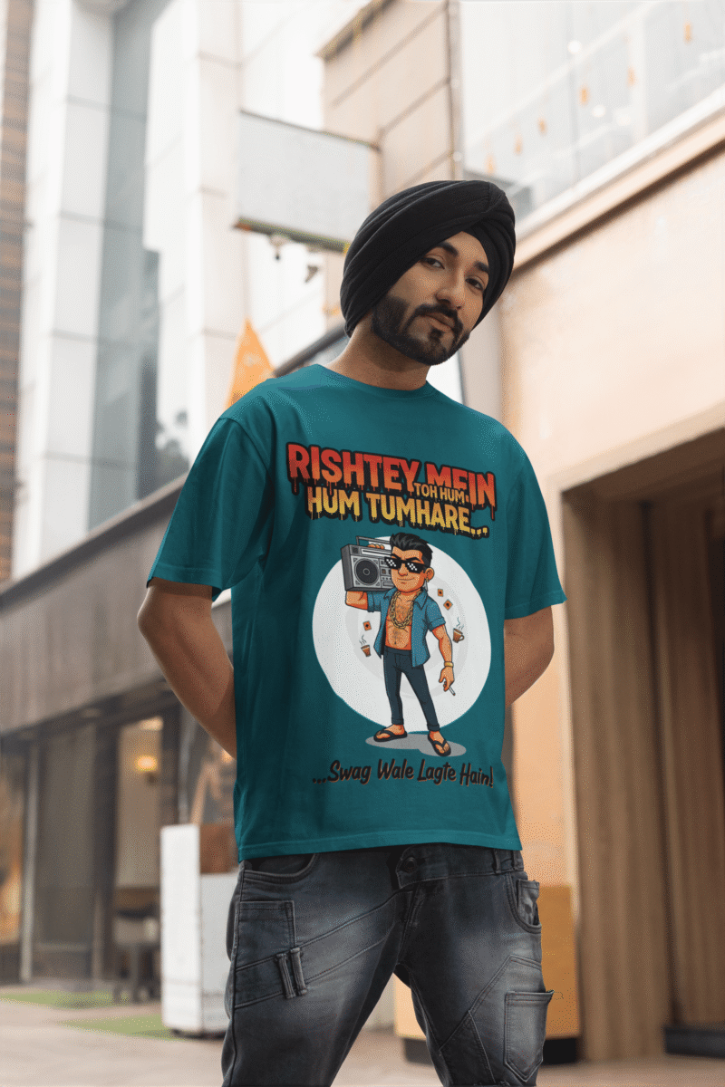 A young man, possibly of Caucasian descent, with short dark hair and a beard, wearing a petrol blue PodLyft Men's Bollywood Swag T-shirt. The t-shirt features a graphic with a funny movie dialogue. He is looking directly at the camera with a confident expression, standing against a plain light grey background.