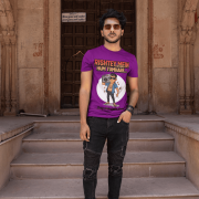A young man, possibly of Caucasian descent, with short dark hair and a beard, wearing a purple PodLyft Men's Bollywood Swag T-shirt. The t-shirt features a graphic with a funny movie dialogue. He is looking directly at the camera with a confident expression, standing against a plain light grey background.