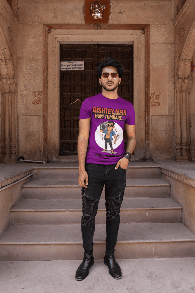 A young man, possibly of Caucasian descent, with short dark hair and a beard, wearing a purple PodLyft Men's Bollywood Swag T-shirt. The t-shirt features a graphic with a funny movie dialogue. He is looking directly at the camera with a confident expression, standing against a plain light grey background.