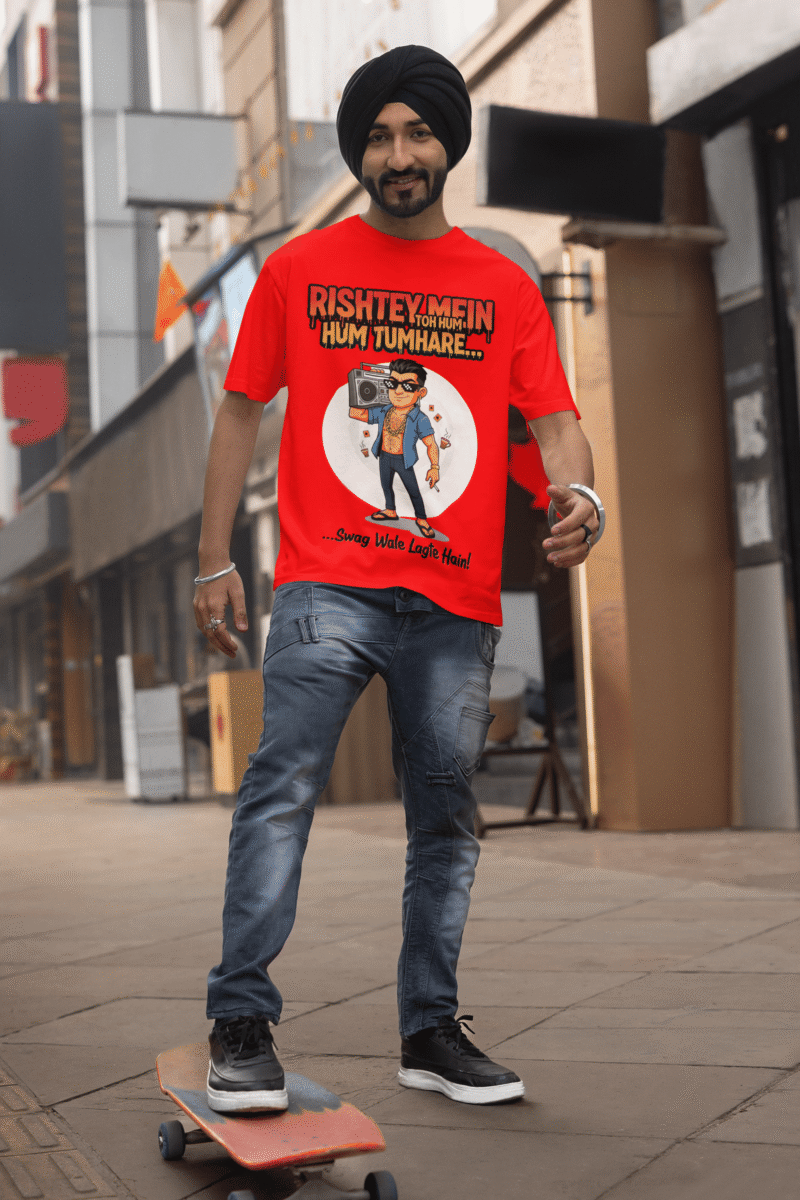 A young man, possibly of Caucasian descent, with short dark hair and a beard, wearing a red PodLyft Men's Bollywood Swag T-shirt. The t-shirt features a graphic with a funny movie dialogue. He is looking directly at the camera with a confident expression, standing against a plain light grey background.