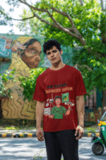 A man wearing a maroon PodLyft Men's Engineering Stress Tee outdoors with a wall mural in the background. The graphic on the shirt shows a stressed-out male engineer surrounded by broken electronics, wires, and coffee, with the Hindi text "MAA KASAM ENGINEER HOON" and a book titled "LEARN PYTHON 7 DAYS!