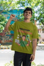 A man smiling and posing in an olive green PodLyft Men's Engineering Stress Tee outdoors with a wall mural in the background. The graphic on the shirt shows a stressed-out male engineer surrounded by broken electronics, wires, and coffee, with the Hindi text "MAA KASAM ENGINEER HOON" and a book titled "LEARN PYTHON 7 DAYS!
