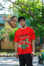 A man smiling and posing in a red PodLyft Men's Engineering Stress Tee outdoors with a wall mural in the background. The graphic on the shirt shows a stressed-out male engineer surrounded by broken electronics, wires, and coffee, with the Hindi text "MAA KASAM ENGINEER HOON" and a book titled "LEARN PYTHON 7 DAYS!
