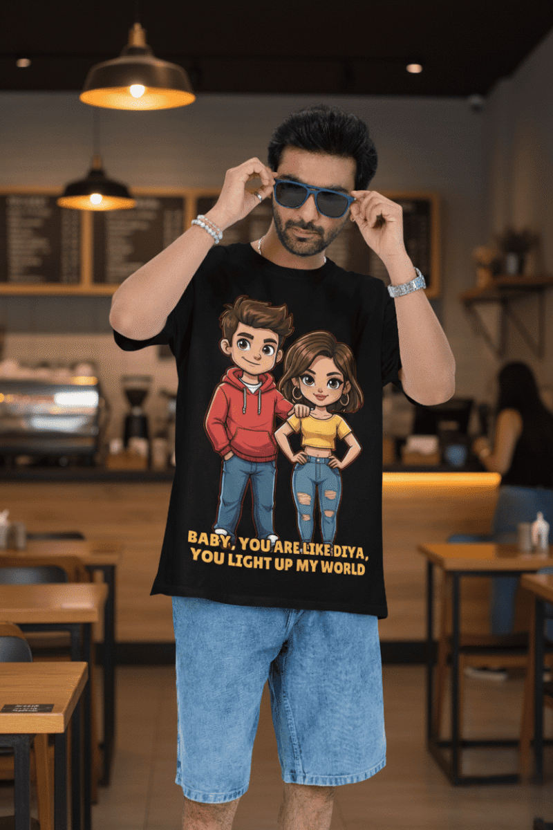 A young man, possibly of Caucasian descent, with short dark hair and a beard, wearing a black PodLyft oversized Light Up T-shirt. The t-shirt features a graphic of a cartoon-style couple and the flirty text, "Baby You Are Like Diya, You Light Up My World." He is looking directly at the camera with a confident expression, standing against a plain light grey background.