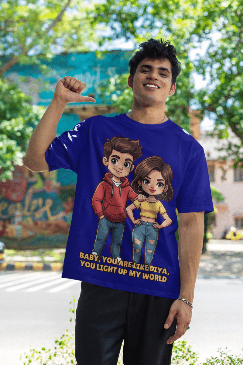 A young man, possibly of Caucasian descent, with short dark hair and a beard, wearing a navy blue PodLyft oversized Light Up T-shirt. The t-shirt features a graphic of a cartoon-style couple and the flirty text, "Baby You Are Like Diya, You Light Up My World." He is looking directly at the camera with a confident expression, standing against a plain light grey background.