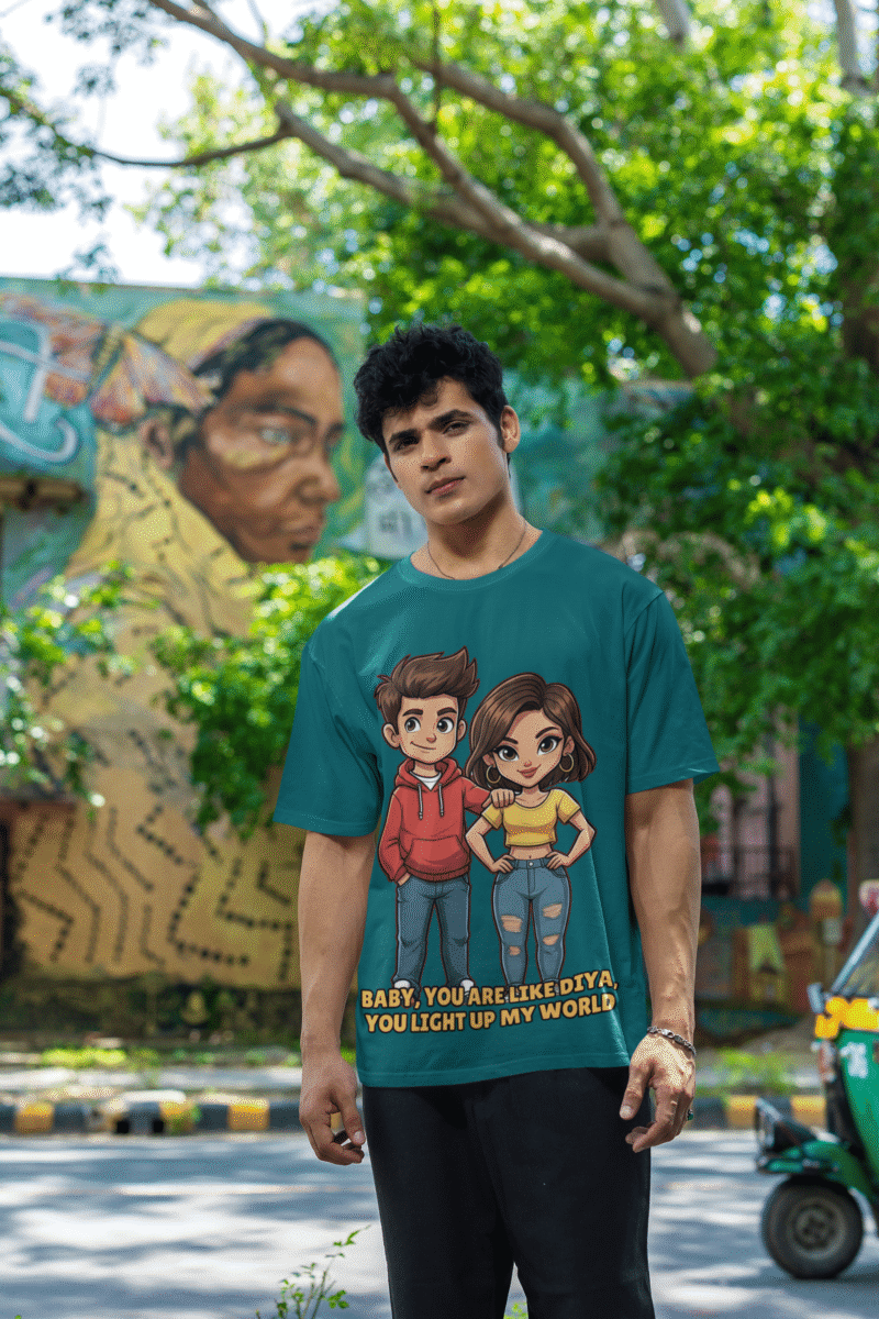 A young man, possibly of Caucasian descent, with short dark hair and a beard, wearing a petrol blue PodLyft oversized Light Up T-shirt. The t-shirt features a graphic of a cartoon-style couple and the flirty text, "Baby You Are Like Diya, You Light Up My World." He is looking directly at the camera with a confident expression, standing against a plain light grey background.