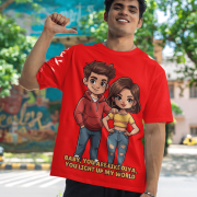 A young man, possibly of Caucasian descent, with short dark hair and a beard, wearing a red PodLyft oversized Light Up T-shirt. The t-shirt features a graphic of a cartoon-style couple and the flirty text, "Baby You Are Like Diya, You Light Up My World." He is looking directly at the camera with a confident expression, standing against a plain light grey background.