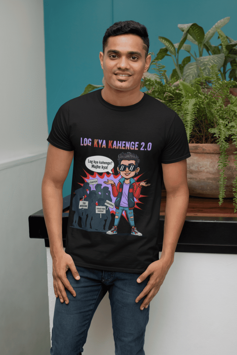 A young man, possibly of Caucasian descent, with short dark hair and a beard, wearing a black PodLyft Log Kya Kahenge T-shirt. The t-shirt features a funny social parody graphic with Hindi text. He is looking directly at the camera with a confident expression, standing against a plain light grey background.