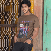 A young man, possibly of Caucasian descent, with short dark hair and a beard, wearing a coffee brown PodLyft Log Kya Kahenge T-shirt. The t-shirt features a funny social parody graphic with Hindi text. He is looking directly at the camera with a confident expression, standing against a plain light grey background.