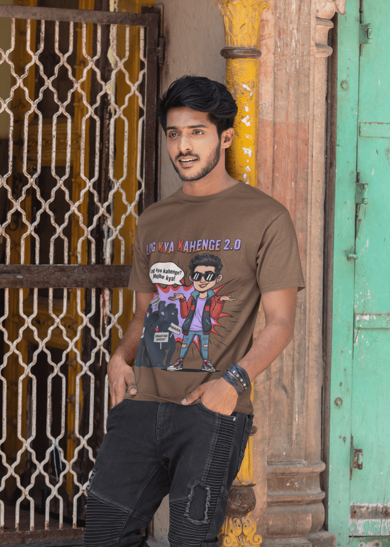 A young man, possibly of Caucasian descent, with short dark hair and a beard, wearing a coffee brown PodLyft Log Kya Kahenge T-shirt. The t-shirt features a funny social parody graphic with Hindi text. He is looking directly at the camera with a confident expression, standing against a plain light grey background.