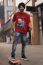 A young man, possibly of Caucasian descent, with short dark hair and a beard, wearing a maroon PodLyft Log Kya Kahenge T-shirt. The t-shirt features a funny social parody graphic with Hindi text. He is looking directly at the camera with a confident expression, standing against a plain light grey background.