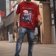 A young man, possibly of Caucasian descent, with short dark hair and a beard, wearing a maroon PodLyft Log Kya Kahenge T-shirt. The t-shirt features a funny social parody graphic with Hindi text. He is looking directly at the camera with a confident expression, standing against a plain light grey background.