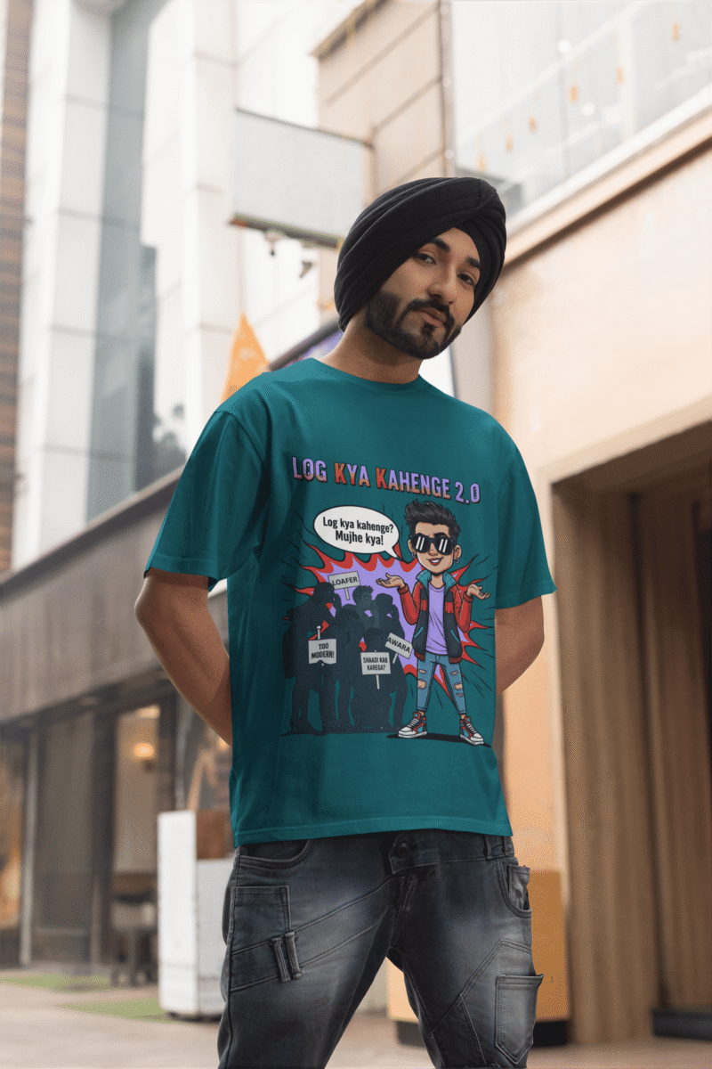 A young man, possibly of Caucasian descent, with short dark hair and a beard, wearing a petrol blue PodLyft Log Kya Kahenge T-shirt. The t-shirt features a funny social parody graphic with Hindi text. He is looking directly at the camera with a confident expression, standing against a plain light grey background.