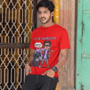 A young man, possibly of Caucasian descent, with short dark hair and a beard, wearing a red PodLyft Log Kya Kahenge T-shirt. The t-shirt features a funny social parody graphic with Hindi text. He is looking directly at the camera with a confident expression, standing against a plain light grey background.