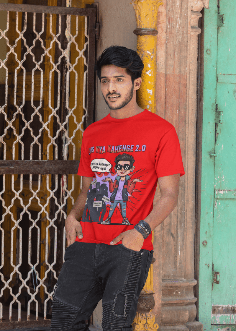 A young man, possibly of Caucasian descent, with short dark hair and a beard, wearing a red PodLyft Log Kya Kahenge T-shirt. The t-shirt features a funny social parody graphic with Hindi text. He is looking directly at the camera with a confident expression, standing against a plain light grey background.