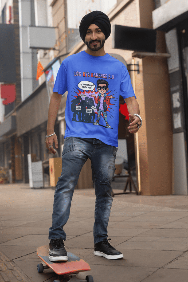 A young man, possibly of Caucasian descent, with short dark hair and a beard, wearing a royal blue PodLyft Log Kya Kahenge T-shirt. The t-shirt features a funny social parody graphic with Hindi text. He is looking directly at the camera with a confident expression, standing against a plain light grey background.