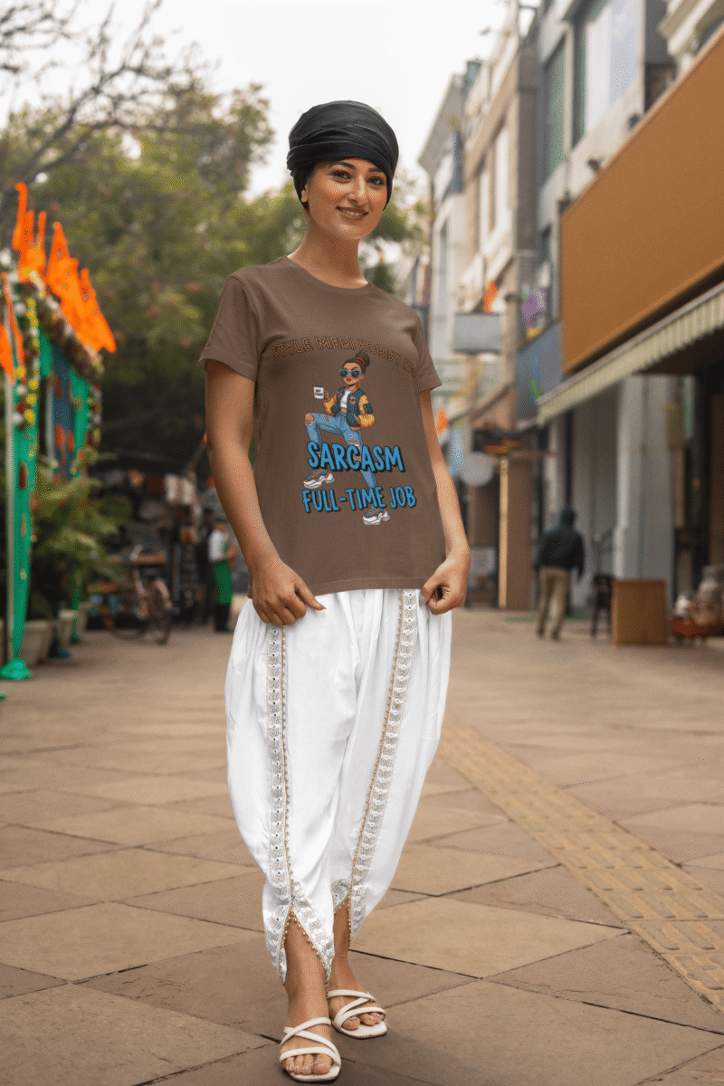 A woman smiling while wearing a coffee brown PodLyft Women's Full-Time Sarcasm Tee and white traditional Indian clothing. She is standing on a city street. The graphic shows a stylized woman holding a coffee cup, with the text "STYLE MERI HOBBY HAI" and "SARCASM FULL-TIME JOB.