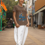 A woman smiling while wearing a coffee brown PodLyft Women's Full-Time Sarcasm Tee and white traditional Indian clothing. She is standing on a city street. The graphic shows a stylized woman holding a coffee cup, with the text "STYLE MERI HOBBY HAI" and "SARCASM FULL-TIME JOB.