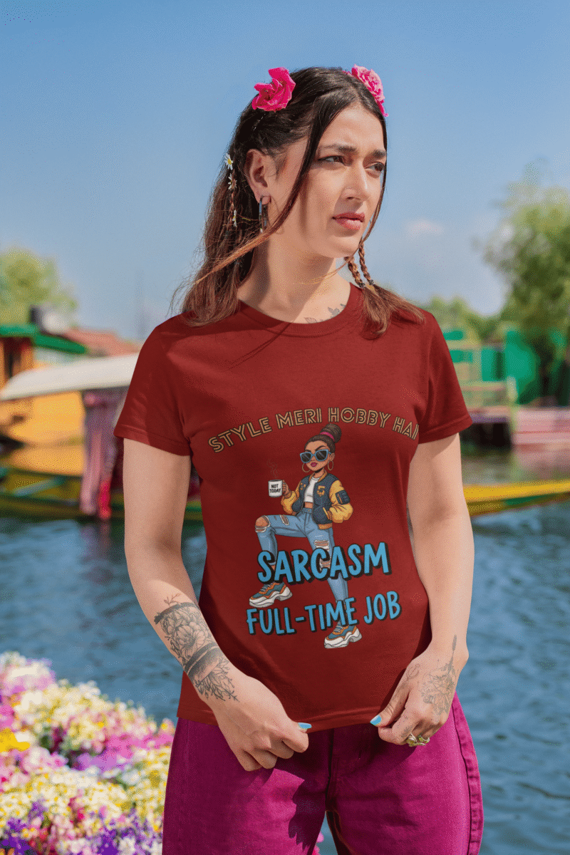 A woman wearing a maroon PodLyft Women's Full-Time Sarcasm Tee outdoors with a lake and mountains in the background. The graphic features a stylized woman holding a coffee cup, with the text "STYLE MERI HOBBY HAI" and "SARCASM FULL-TIME JOB.
