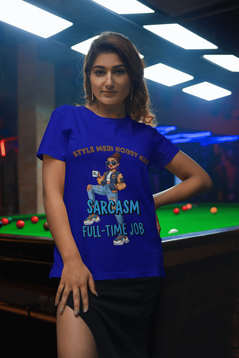 A woman in a navy blue PodLyft Women's Full-Time Sarcasm Tee standing next to a pool table indoors. The graphic features a stylized woman holding a coffee cup, with the text "STYLE MERI HOBBY HAI" and "SARCASM FULL-TIME JOB.