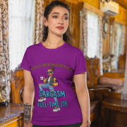 A woman wearing a purple PodLyft Women's Full-Time Sarcasm Tee in a cozy, wood-paneled room. The graphic features a stylized woman holding a coffee cup, with the text "STYLE MERI HOBBY HAI" and "SARCASM FULL-TIME JOB.