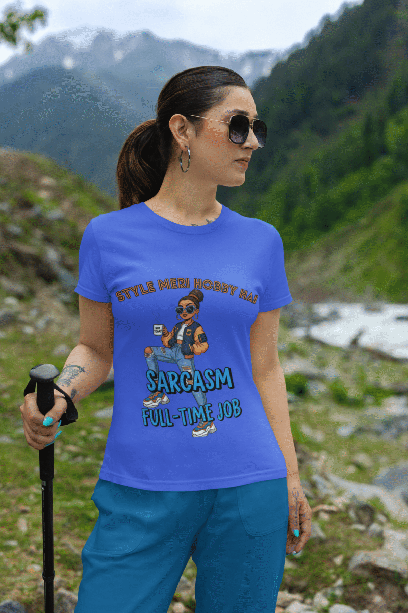 A woman wearing a royal blue PodLyft Women's Full-Time Sarcasm Tee in a mountain setting, holding a hiking pole. The graphic features a stylized woman holding a coffee cup, with the text "STYLE MERI HOBBY HAI" and "SARCASM FULL-TIME JOB.