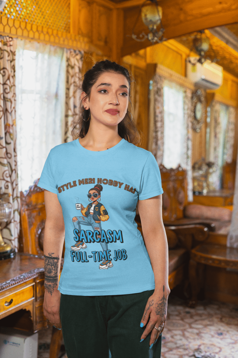 A woman wearing a sky blue PodLyft Women's Full-Time Sarcasm Tee in a cozy, wood-paneled room. The graphic features a stylized woman holding a coffee cup, with the text "STYLE MERI HOBBY HAI" and "SARCASM FULL-TIME JOB."