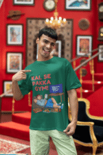 A man winking and pointing to his bottle green PodLyft Men's Procrastinator Gym Tee in a room with a red wall and staircase. The graphic shows a cartoon man relaxing on a couch surrounded by junk food, with the Hindi text "KAL SE PAKKA GYM!" and a TV screen saying "MOTIVATIONAL WORKOUT: 4 HOURS LEFT TO START.