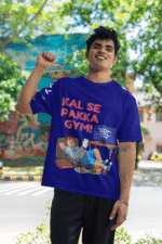 A man smiling and posing in a navy blue PodLyft Men's Procrastinator Gym Tee outdoors with a wall mural in the background. The graphic shows a cartoon man relaxing on a couch surrounded by junk food, with the Hindi text "KAL SE PAKKA GYM!" and a TV screen saying "MOTIVATIONAL WORKOUT: 4 HOURS LEFT TO START.