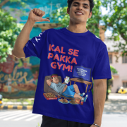 A man smiling and posing in a navy blue PodLyft Men's Procrastinator Gym Tee outdoors with a wall mural in the background. The graphic shows a cartoon man relaxing on a couch surrounded by junk food, with the Hindi text "KAL SE PAKKA GYM!" and a TV screen saying "MOTIVATIONAL WORKOUT: 4 HOURS LEFT TO START.