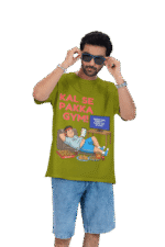 A man wearing an olive green PodLyft Men's Procrastinator Gym Tee against a solid dark green background, adjusting his blue sunglasses. The graphic shows a cartoon man relaxing on a couch surrounded by junk food, with the Hindi text "KAL SE PAKKA GYM!" (Definitely Gym From Tomorrow!) and a TV screen saying "MOTIVATIONAL WORKOUT: 4 HOURS LEFT TO START.