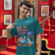 A man winking and pointing to his petrol blue PodLyft Men's Procrastinator Gym Tee in a room with a red wall and staircase. The graphic shows a cartoon man relaxing on a couch surrounded by junk food, with the Hindi text "KAL SE PAKKA GYM!" and a TV screen saying "MOTIVATIONAL WORKOUT: 4 HOURS LEFT TO START.