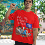 A man smiling and posing in a red PodLyft Men's Procrastinator Gym Tee outdoors with a wall mural in the background. The graphic shows a cartoon man relaxing on a couch surrounded by junk food, with the Hindi text "KAL SE PAKKA GYM!" and a TV screen saying "MOTIVATIONAL WORKOUT: 4 HOURS LEFT TO START.