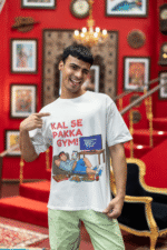 A man winking and pointing to his white PodLyft Men's Procrastinator Gym Tee in a room with a red wall and staircase. The graphic shows a cartoon man relaxing on a couch surrounded by junk food, with the Hindi text "KAL SE PAKKA GYM!" and a TV screen saying "MOTIVATIONAL WORKOUT: 4 HOURS LEFT TO START.